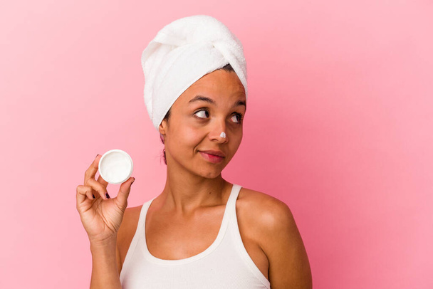 Joven mujer de raza mixta sosteniendo crema facial aislada sobre fondo rosa soñando con lograr objetivos y propósitos - Foto, imagen