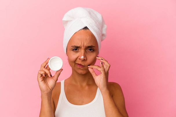 Young mixed race woman holding facial cream isolated on pink background with fingers on lips keeping a secret. - Fotó, kép