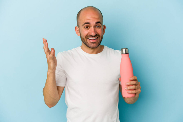 Young bald man holding canteen isolated on blue background  receiving a pleasant surprise, excited and raising hands. - Photo, Image