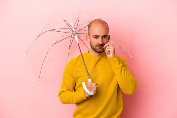 Молодой кавказский лысый мужчина держит зонтик изолированный на розовом фоне с пальцами на губах, хранящими секрет. - Фото, изображение