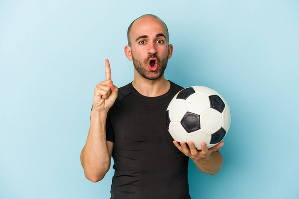 Junger Geschäftsmann mit Glatze, der isoliert auf blauem Hintergrund Fußball spielt, hat eine großartige Idee, ein Konzept der Kreativität. - Foto, Bild