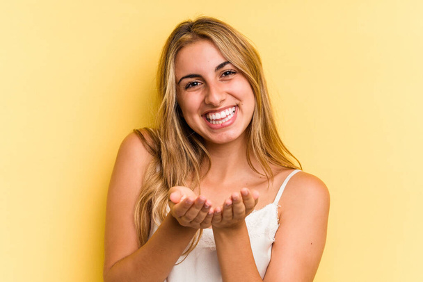 Young caucasian blonde woman isolated on yellow background  holding something with palms, offering to camera. - Photo, Image