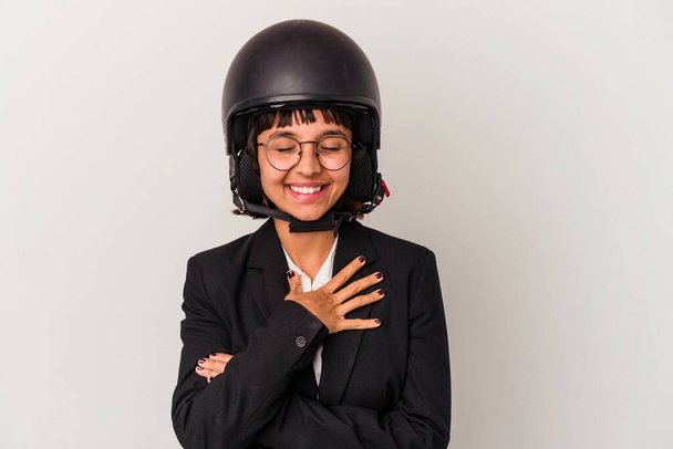 Joven mujer de negocios de raza mixta que lleva un casco de moto aislado se ríe en voz alta manteniendo la mano en el pecho. - Foto, imagen