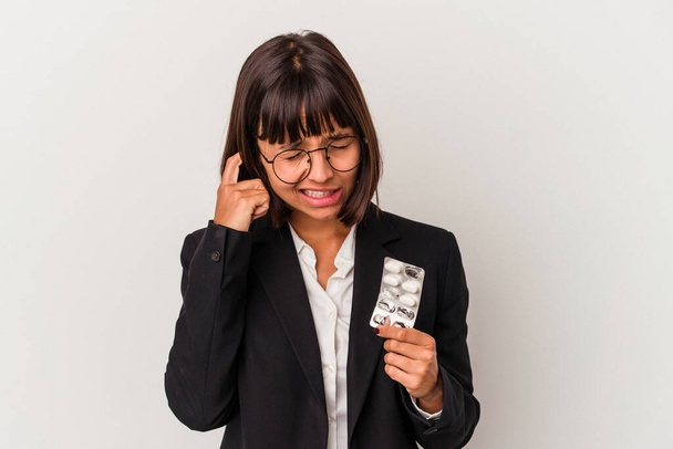 Молодая деловая женщина смешанной расы держит таблетки изолированные закрывающие уши руками. - Фото, изображение