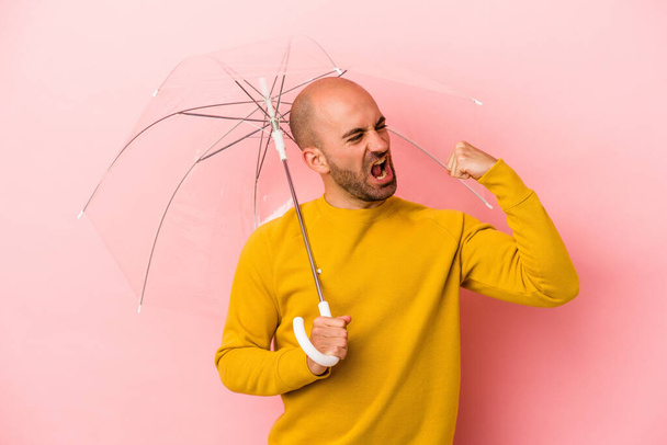 Giovane uomo calvo caucasico che tiene l'ombrello isolato su sfondo rosa alzando il pugno dopo una vittoria, concetto vincitore. - Foto, immagini