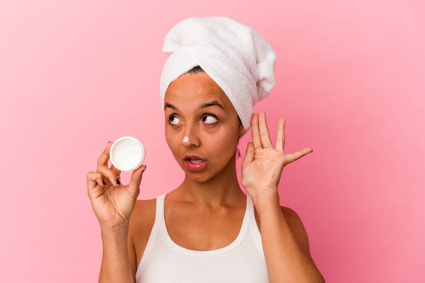 Young mixed race woman holding facial cream isolated on pink background trying to listening a gossip. - Photo, Image