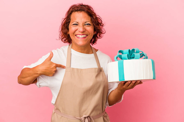 Středního věku pečivo latinsky žena drží dort izolované na růžovém pozadí osoba ukazuje ručně na košili kopírovat prostor, hrdý a sebejistý - Fotografie, Obrázek
