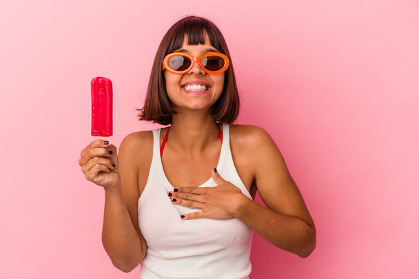 Молодая женщина смешанной расы, держащая мороженое на голубом фоне, громко смеется, держа руку на груди.. - Фото, изображение