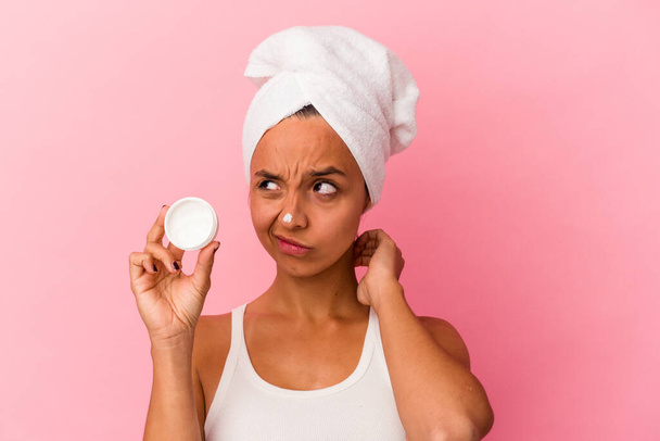 Young mixed race woman holding facial cream isolated on pink background touching back of head, thinking and making a choice. - Foto, imagen