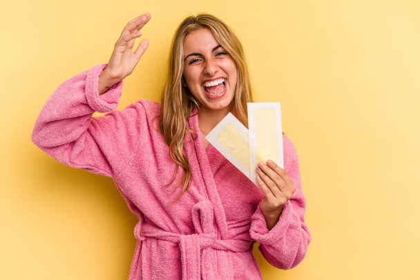 Young caucasian blonde woman wearing bathrobe holding depilatory bands isolated on yellow background  receiving a pleasant surprise, excited and raising hands. - Valokuva, kuva