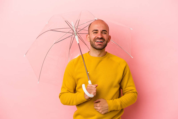 Young caucasian bald man holding umbrella isolated on pink background  laughing and having fun. - Photo, Image