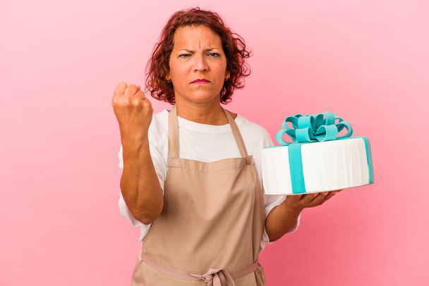 Middle age pastry latin woman holding a cake isolated on pink background showing fist to camera, aggressive facial expression. - Fotoğraf, Görsel