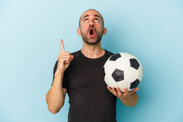 Young business bald man playing football isolated on blue background  pointing upside with opened mouth. - Фото, изображение