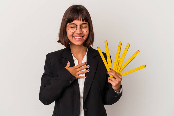 Young mixed race business woman holding a measure tape isolated on white background laughs out loudly keeping hand on chest. - Foto, immagini