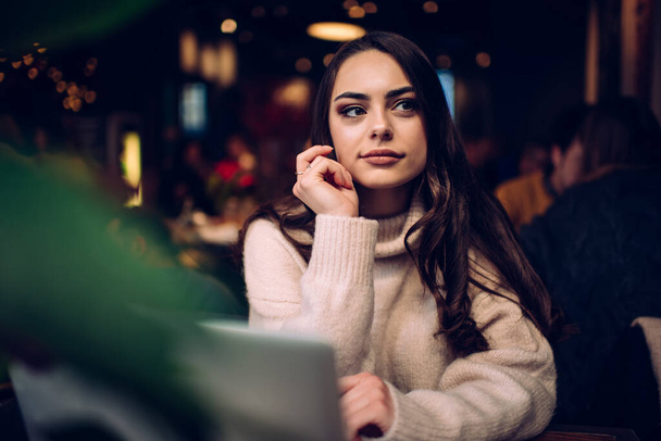 Pensive young woman with long hair in casual sweater sitting and touching face while looking thoughtfully away in contemporary workspace - Fotoğraf, Görsel
