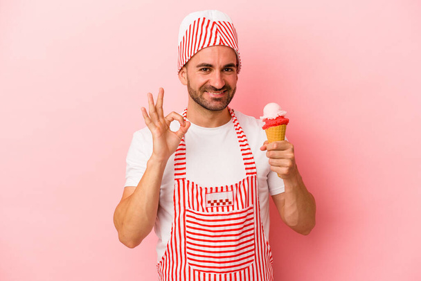 若いですアイスクリームメーカー男保持アイスクリーム隔離オンピンクの背景陽気で自信を持ってショーokジェスチャー. - 写真・画像