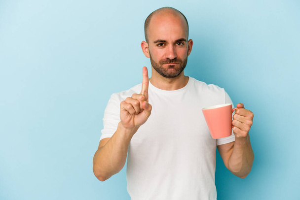 Young bald man holding a mug isolated on blue background  showing number one with finger. - Photo, image