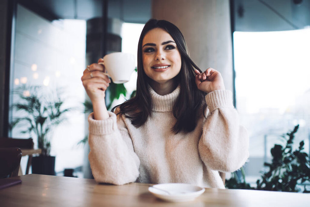 Happy female in casual wear smiling and looking away while drinking cup of hot coffee and spending time in cafe - Фото, зображення