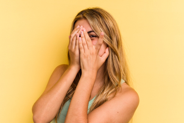 Young caucasian blonde woman isolated on yellow background  blink at the camera through fingers, embarrassed covering face. - Photo, Image