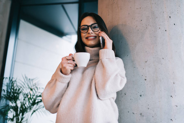 Low angle of happy female in cashmere sweater drinking coffee while communicating on cellphone and looking at camera with smile - Φωτογραφία, εικόνα