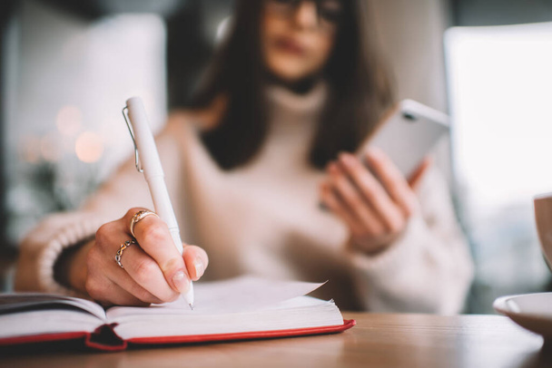 Blurred female freelancer in casual wear and glasses sitting at wooden table and messaging on smartphone while writing ideas in copybook in cafe - Foto, afbeelding
