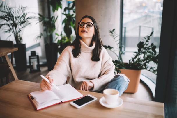 High angle of thoughtful female freelancer in casual wear and glasses sitting at wooden table in cafe and taking notes in copybook and looking away - Фото, изображение
