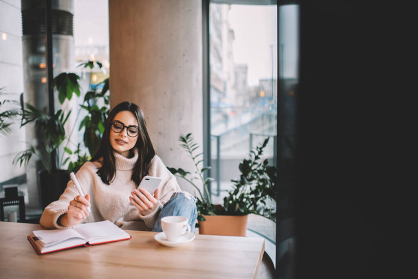 Serious female freelancer in casual wear and eyeglasses sitting at wooden table and browsing smartphone while taking notes in copybook in cafe - Foto, Imagem