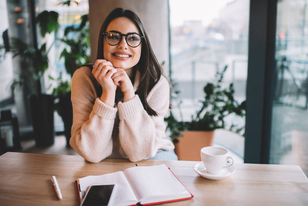 Cheerful female in casual sweater and eyeglasses sitting at wooden table in cozy cafe and looking away while enjoying weekend - Фото, зображення