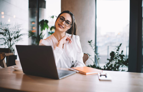 Cheerful female freelancer in casual outfit and eyewear sitting at wooden table and browsing laptop while spending time in cafe and looking at camera - Foto, Imagen