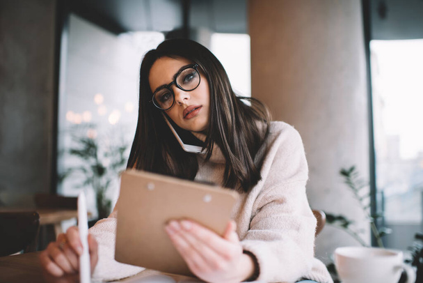 Low angle of thoughtful female freelancer in casual outfit and glasses sitting in modern cafe with clipboard in hand and calling on smartphone while looking at camera - Fotoğraf, Görsel