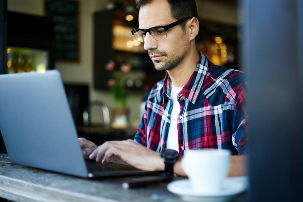 Crop konzentriert männlich in lässigem Outfit und Brille sitzt am Holztisch und arbeitet an Remote-Projekt auf Laptop im Café - Foto, Bild