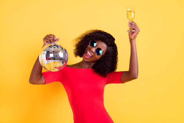 Photo of young beautiful charming excited afro girl hold disco ball drinking champagne isolated on yellow color background - Фото, зображення