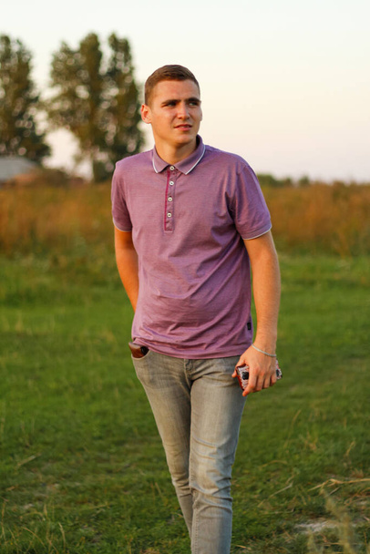 Счастливые люди. Портрет серьезного молодого брюнетки в фиолетовой рубашке поло, стоящего на открытом воздухе на фоне природы в летний день. Парень 20 лет. Тысячелетнее поколение. Снаружи. Серьезный человек. В целом. - Фото, изображение