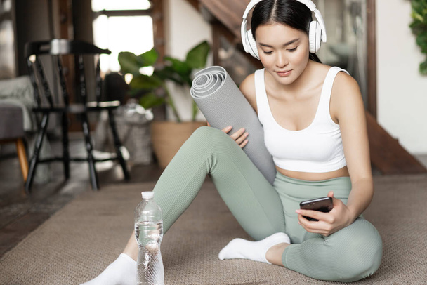 Νεαρή Ασιάτισσα με μπουκάλι νερό, μουσική ακρόασης ή podcast σε ακουστικά κατά τη διάρκεια της γυμναστικής στο σπίτι, κάθεται στο πάτωμα πριν από την άσκηση - Φωτογραφία, εικόνα