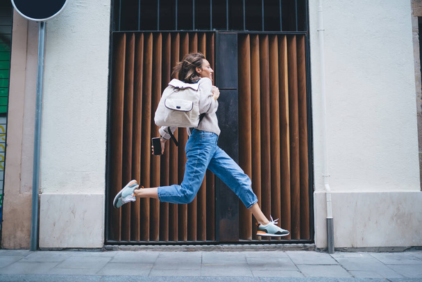 Side view of stylish focused woman in denim and sneakers with backpack being late and running rapidly along pavement against concrete building - Fotoğraf, Görsel