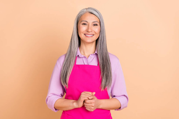 Photo of cute white hairdo aged lady hold hands wear purple shirt pink apron isolated on beige color background - Foto, Imagem