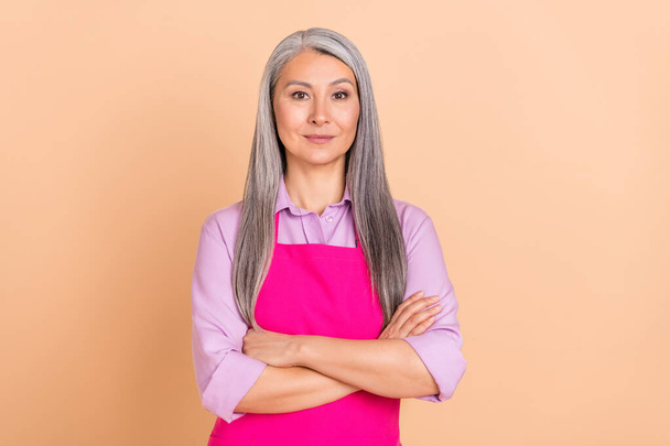 Photo of strict grey hairdo aged lady crossed arms wear purple shirt pink apron isolated on beige color background - Фото, зображення