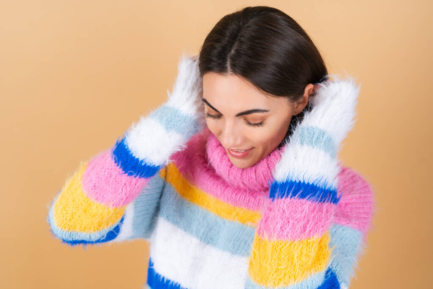 Молодая женщина на бежевом фоне в ярком разноцветном уютном вязаном свитере мило улыбается - Фото, изображение