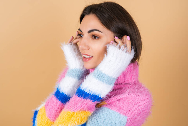 Молодая женщина на бежевом фоне в ярком разноцветном уютном вязаном свитере мило улыбается - Фото, изображение