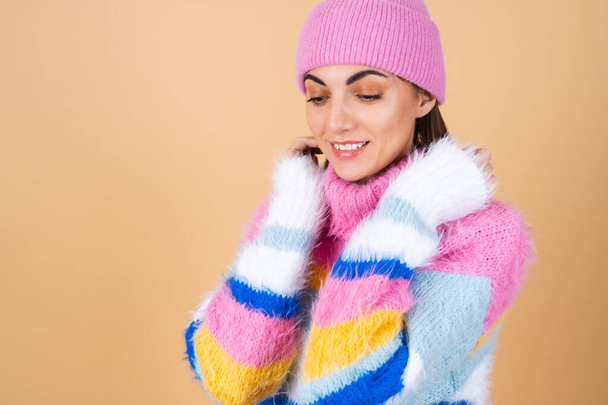 Mujer joven sobre un fondo beige en un suéter de punto acogedor multicolor brillante y un sombrero se abraza cómodamente, calidez y comodidad - Foto, imagen