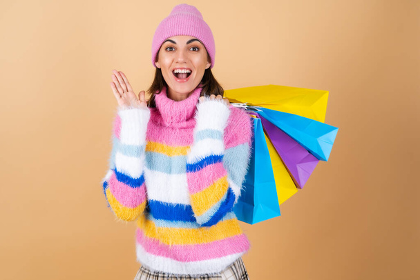 Bej bir arka planda parlak renkli örgü süveter giyen genç bir kadın, ekose bir etek alışveriş paketlerini heyecanla haykırıyor - Fotoğraf, Görsel