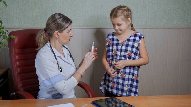 Een vrouwelijke arts in een witte jas legt uit aan een meisje dat gevaccineerd zal worden - Foto, afbeelding