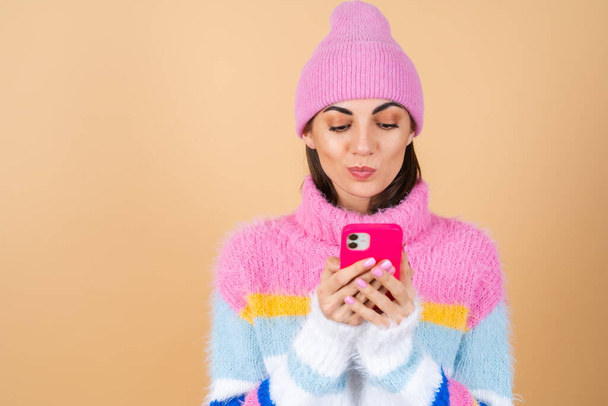 Mujer joven sobre un fondo beige en un suéter de punto y un sombrero lindo cuidadosamente escribiendo un mensaje en el teléfono, mordiéndose el labio - Foto, Imagen