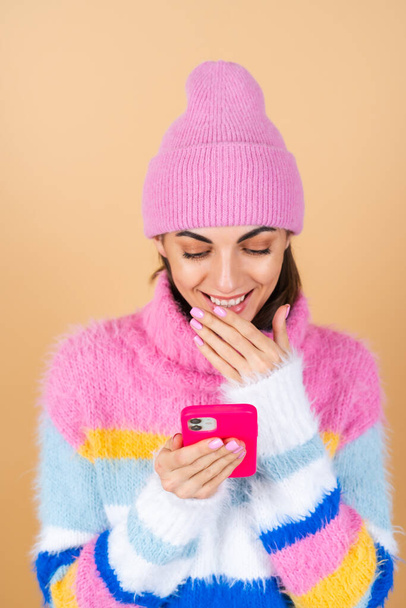 Молодая женщина на бежевом фоне в вязаном свитере и шляпе с мобильным телефоном смеется с удивлением и весельем, читает сообщения, шутит, поздравляет - Фото, изображение