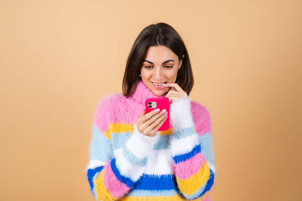Młoda kobieta na beżowym tle w dzianinie sweter z telefonem patrzy na ekran troskliwie ładny gryzie jej palec - Zdjęcie, obraz