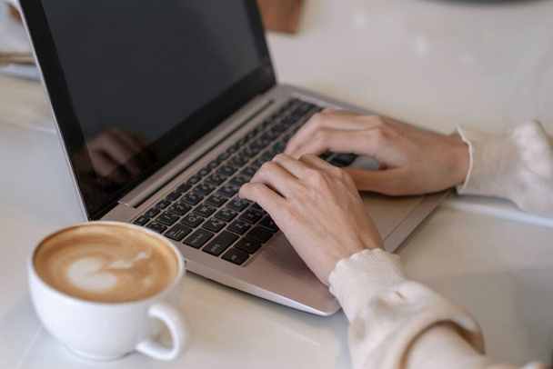 Bezárt nő kéz a fehér pulóver gépelés laptopján elmosódott fehér kávé csésze az asztalon, woring kávézóban vagy munkaterület koncepció. - Fotó, kép