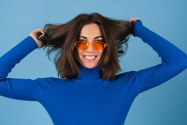 Молодая женщина в гольф на голубом фоне в стильных ярко-оранжевых очках позирует в хорошем настроении - Фото, изображение