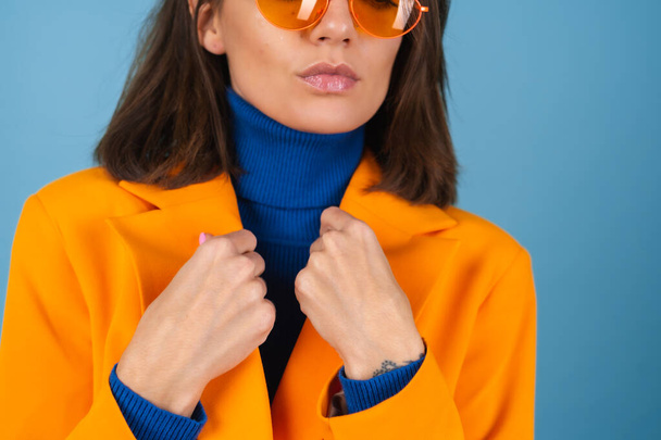 膝丈でオーバーサイズのジャケットを着たファッショナブルな若い女性に青の背景にスタイリッシュな明るいオレンジ色のメガネポーズ - 写真・画像