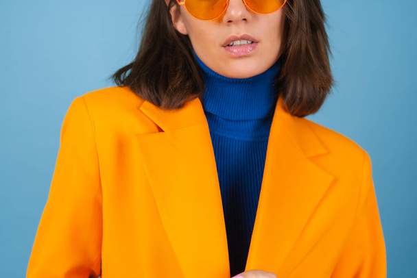 Mujer joven de moda en una chaqueta de gran tamaño y longitud de rodilla sobre un fondo azul en elegantes gafas de color naranja brillante posando - Foto, imagen