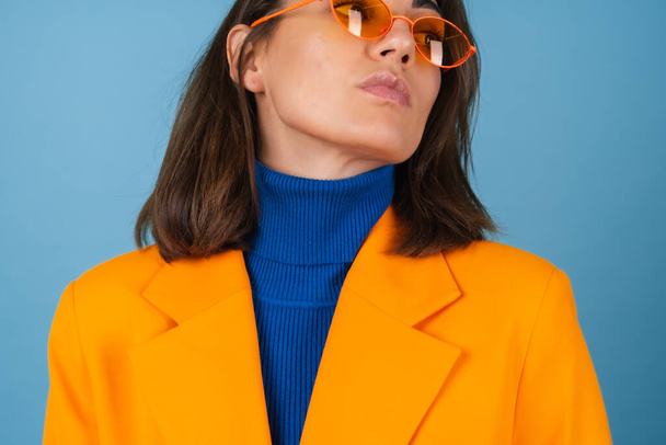Mujer joven de moda en una chaqueta de gran tamaño y longitud de rodilla sobre un fondo azul en elegantes gafas de color naranja brillante posando - Foto, imagen